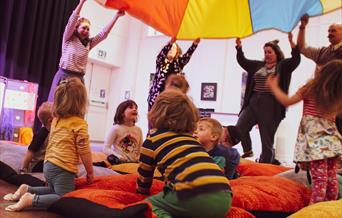 Bright photograph of children sitting on fluffy cushions under a bit coloured parachute.