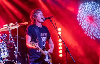 Singer Alex Lipinski playing guitar and singing into a microphone while performing a concert on stage at the Grand Pier, Weston-super-Mare