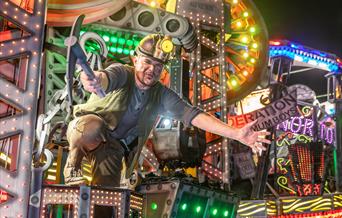 A performer in a miner's helmet and with a mining pick axe on board a brightly coloured illuminated float at the Weston-super-Mare Carnival