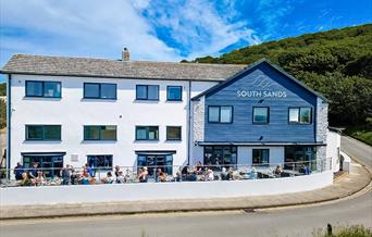 Exterior of white hotel front with patrons dining in the front, a road winding round it and woods in the background