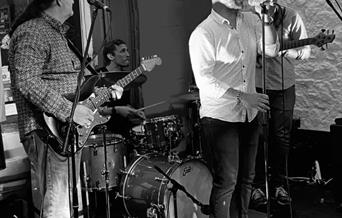 Black and white picture of three band members performing in front of their seated drummer