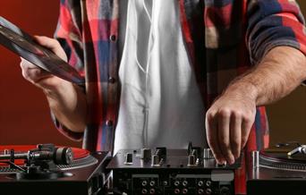 A man in a checked shirt and white undershirt (cropped without a head) holding a record above some decks