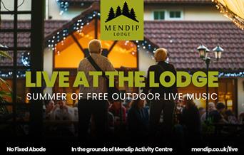 Two musicians (man and woman) playing guitar and singing into microphone under tented woodland canopy. People sat on picnic benches in front of Mendip