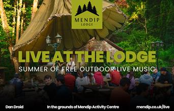 Two musicians (man and woman) playing guitar and singing into microphone under tented woodland canopy. People sat on picnic benches in front of Mendip