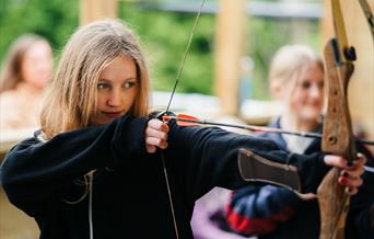 Teenage girl with blonde hair, wearing a black hoodie, pulling back an arrow on an archery bow. Looking straight down the arrow with determination.