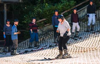 Middle-aged lady wearing skis, gloves and a helmet, looking forward as she glides along the bottom of the ski slope. In the background a Mendip Activi