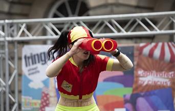 A lady in a red and yellow lifeguard costume staring through an over-large pair of binoculars