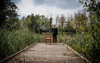 A young person stands by a chair at the end of a wooden walkway. There are trees and plants around them