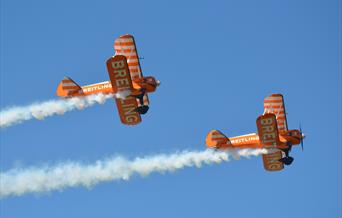 Two orange bi-planes with white plumes of smoke flying across a blue sky