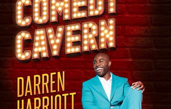 Comedian Darren Herriott wearing a light blue suit sitting casually on the floor in front of a brick wall with the words Comedy Cavern