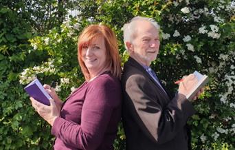 Two people standing back-to-back holding books. The background is a bush with white flowers.