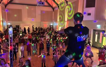 The back of a performer on stage entertaining a crowd of young children and families on a dancefloor