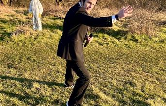 A man in a dark suit impersonating James Bond while in the background is someone in a white police crime scene suit