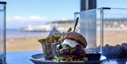 A platter of burger and chips in an open window with the view of a beach with a pier and town in the background