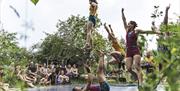 A man laying on his back on a trampoline with hands and legs reaching to the sky, while other perfomers jump around on the trampoline. A crowd watches