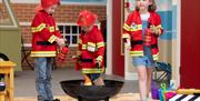 Three young children dressed up as firemen standing around a firepit in a model village