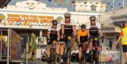 A group of four cyclists riding under the Grand Pier sign in Weston-super-Mare as they ride up the boardwalk at the finish of the Chase The Sun bike r