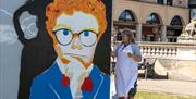 A female street artist at work to the side of a mural of a  man wearing glasses and looking thoughtful