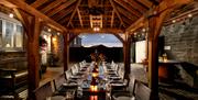 A large dining area under wooden beams which opens out to the evening sky. The large table at this Webbington Farm Holiday cottage is lit with lanters