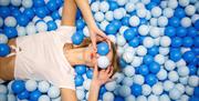 A lady lying in a children's ball pit filled with blue and white balls. The lady is holding two of the balls over  her eyes