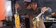 A lady stall holder at the Eat Weston Food Festival holding a tong full of noodles and another full of vegetables over two hot food platters