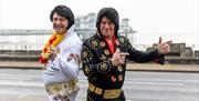 Two Elvis impersonators dressed in iconic Elvis suits, one in the white and one in the black, standing back to back and looking at the camera as they