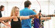 A lady with blonde hair pulls back the bow and is about to fire during an archery lesson at the Mendip Activity Centre. In the background the instruct