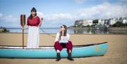Two women on a beach in a blue canoe. One is sitting with a typewriter, smiling at the other who is standing with a paddle in her hands. The canoe sit
