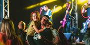 A dad lifting his daughter at the Mendip Family Festival with a band on stage in the background during a night time concert