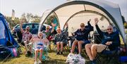 A family sitting outside their tent and waving to the camera at the Mendip Basecamp, near Weston-super-Mare