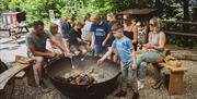 A group of adults and children cooking food on skewers in a firepit at the Mendip Activity Centre, near Weston-super-Mare