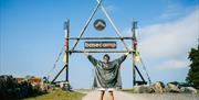 A person standing with their arms out wide under the main Mendip Basecamp sign, near Weston-super-Mare