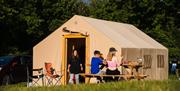 A family sitting on a table and eating outside a large framed tent with a pitched roof and wooden door frame at The Mendip Basecamp, near Weston-super