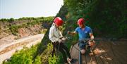 A teenage girl standing on a wooden platform on a rockface being given instructions before her abseil by an instructor at The Mendip Activity Centre