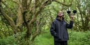 Man outdoors, holding up sound equipement. He is looking to the sky with trees above him