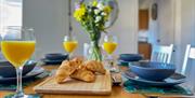 A wooden breakfast table set with glasses of orange juice and croissants and a blue dining service at the Norton Court Farm self-catering holiday cott