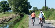 Three cyclists riding down a country lane with a river by the side of it