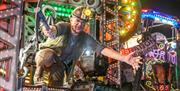 A performer in a miner's helmet and with a mining pick axe on board a brightly coloured illuminated float at the Weston-super-Mare Carnival