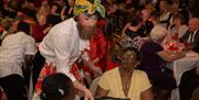 Man with beard and colourful headscarf, with make up on, serving wine to two women at a table in a room full of people, also at tables