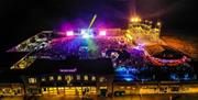 Aerial night time view of an entertainment venue with a funfair, bright stage lights and crowds of people