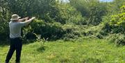 Nature Recovery Ranger, Nathan Orr, in a sun hat, gestures towards dense greenery in a sunny meadow clearing - brambles and trees in the foreground.