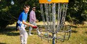 Boy in a blue shirt smiling as he throws a frisbee into a disc golf basket, with a man in sunglasses behind him. The basket is marked with the number