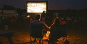 Outdoor cinema screen with audience sat in the dark. Couple sat on camp chairs looking at screen. Glow of a campfire.
