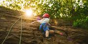 Child wearing a helmet and harness rock climbing at Mendip Activity Centre with the sun shining through the trees.