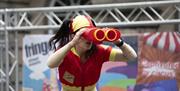 A lady in a red and yellow lifeguard costume staring through an over-large pair of binoculars