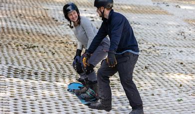 A smiling lady in a helmet on a snowboard receiving tuition from an instructor at the Mendip Activity Centre