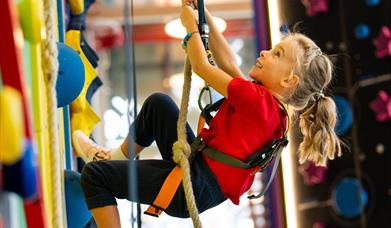 A young blonde girl in a red t-short and black trousers holding onto a rope as she climbs a colourful climbing wall