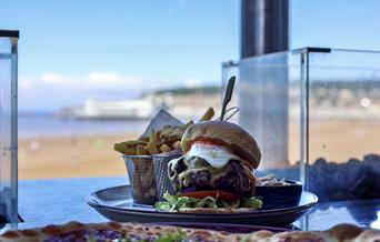 A platter of burger and chips in an open window with the view of a beach with a pier and town in the background