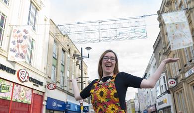 Woman with arms in air on Weston high street with two flags hanging behind her under the welcome to weston sign