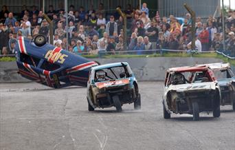 A banger racing car on its roof with three other three-wheeler cars in front